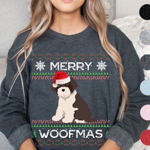 May include: Dark gray sweatshirt with a festive design. The sweatshirt features a graphic of a dog wearing a Santa hat, with the words "MERRY WOOFMAS" in white. The design includes snowflakes, candy canes, and Christmas trees.
