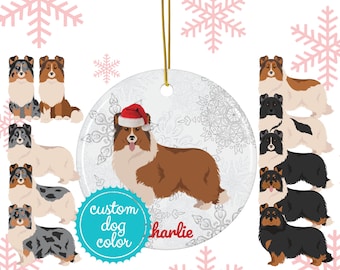 Op de afbeelding: Ronde keramische kerstversiering met een Sheltie-hond met een kerstmuts, met de naam "Charlie" in rood schrift. De versiering heeft een gouden touwtje om op te hangen en is omgeven door sneeuwvlokontwerpen. De woorden "custom dog color" staan op een blauwgroene badge.