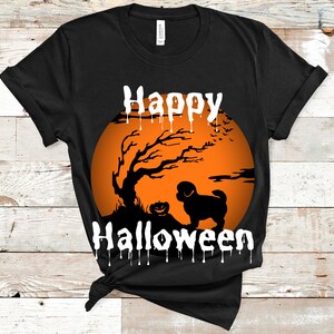 May include: A black short-sleeved t-shirt featuring a graphic of an orange full moon, a silhouette of a tree, a small pumpkin, and a dog. The text "Happy Halloween" is printed in white with a dripping effect above and below the graphic.