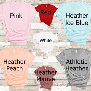 May include: A selection of t-shirts in various colors, including pink, red, white, heather ice blue, heather peach, heather mauve, and athletic heather. The shirts are all folded and laid out on a wooden surface.
