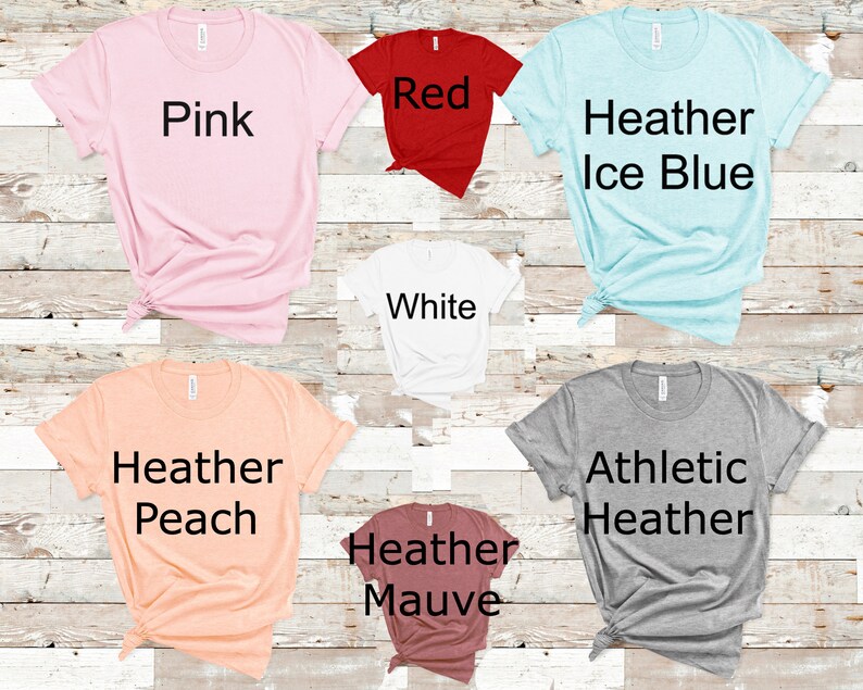 May include: A selection of t-shirts in various colors, including pink, red, white, heather ice blue, heather peach, heather mauve, and athletic heather. The shirts are all folded and laid out on a wooden surface.