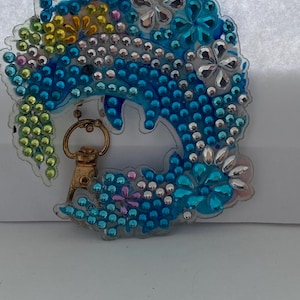 Puede incluir: Un llavero transparente en forma de ola adornado con pedrería azul, plateada y colorida. El diseño incluye adornos en forma de flor y un cierre dorado. El llavero es un accesorio decorativo.