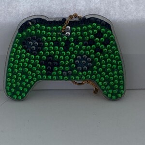 Puede incluir: Un llavero de mando de videojuego verde y negro cubierto de pedrería. El mando tiene un contorno plateado y un cierre y cadena dorados. El mando está cubierto de pedrería verde y negra.