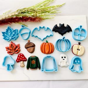 May include: A collection of light blue plastic cookie cutters and their corresponding finished biscuits, including shapes of leaves, acorns, bats, pumpkins, apples, mushrooms, jumpers, and ghosts, arranged in rows on a white rectangular board.