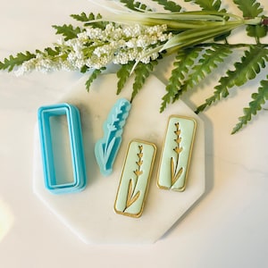 May include: A set of three cookie cutters, two are in the shape of a rectangle with a lavender design and one is a single lavender design. The cookie cutters are on a white surface with a green plant in the background.
