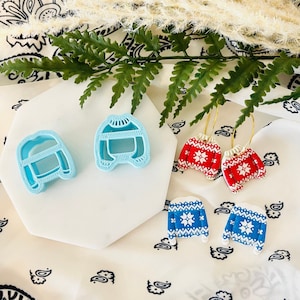 Könnte beinhalten: Ein Set mit vier Ohrringen zum Thema Weihnachten. Zwei Paar Ohrringe zeigen ein rotes und weißes Pullover-Design, und zwei Paar zeigen ein blaues und weißes Pullover-Design. Die Ohrringe werden auf einer weißen Oberfläche mit einem schwarz-weißen gemusterten Stoff präsentiert.