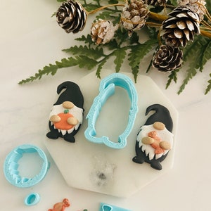 May include: A set of blue plastic cookie cutters featuring a gnome, pumpkin, candy, and a smaller gnome cutter. There are also two small fondant pieces, one orange and one brown, shaped like a pumpkin and candy.