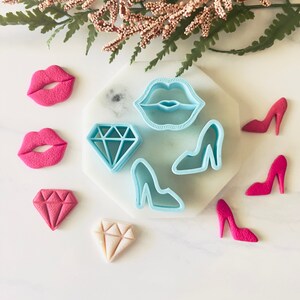 May include: A set of blue plastic cookie cutters in the shapes of lips, a diamond, and a high heel shoe. There are also pink and red fondant cookies in the shapes of lips, a diamond, and high heel shoes.