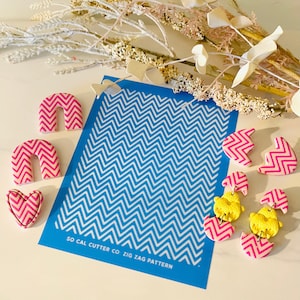 May include: A blue plastic stencil with a white zig zag pattern. The stencil is labeled "SO CAL CUTTER CO. ZIG ZAG PATTERN". There are several pink and white clay earrings with a zig zag pattern on a white surface.