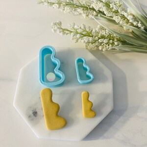 Pode incluir: Dois cortadores de biscoitos de plástico azul em forma de uma forma ondulada e irregular. Também são mostrados dois biscoitos, feitos com os cortadores de biscoitos, de cor amarelo claro.