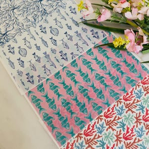 May include: A collection of patterned paper sheets featuring marine life and coral designs. The patterns include blue coral, fish in various shades of blue, and turquoise fish on a pink background. The sheets are arranged in a layered fashion.