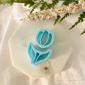 May include: A light blue plastic cookie cutter shaped like a tulip flower. The cutter is on a white marble surface.