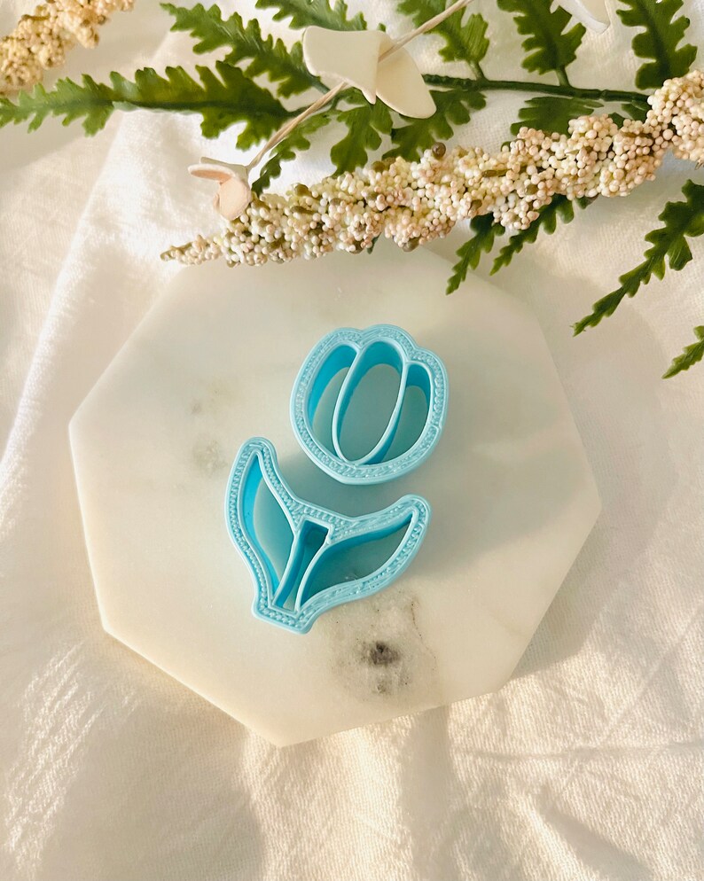 May include: A light blue plastic cookie cutter shaped like a tulip flower. The cutter is on a white marble surface.