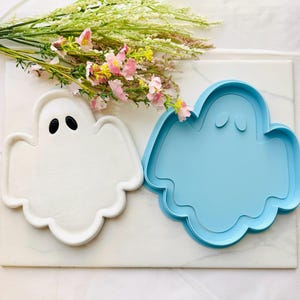 May include: A white ghost-shaped ceramic tray with two black oval eyes and a matching light blue plastic ghost-shaped cookie cutter. The items are arranged together, with a cluster of pink and white flowers positioned above them.
