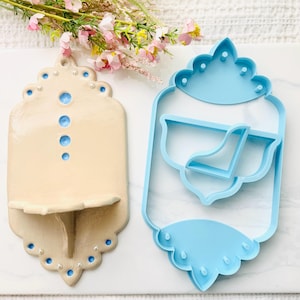 May include: A beige ceramic wall shelf with blue dot accents and a matching light blue plastic cookie cutter. The shelf features a scalloped edge and a small protruding ledge. The cookie cutter mirrors the shape of the shelf with internal dividers.