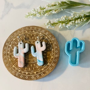 May include: A pair of handmade cactus earrings with a pink and blue marbled design. The earrings are displayed on a wooden tray with a geometric pattern. A blue plastic cactus mold is also shown.