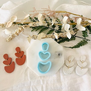 May include: A set of three blue polymer clay cutters in the shape of a heart, a crescent moon, and a rounded rectangle. The cutters are on a white marble surface. There are two pairs of earrings on a white fabric background. One pair is made of brown clay and the other is made of white clay with black speckles.