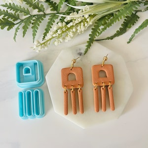 May include: A pair of terracotta-colored clay earrings with a geometric design. Each earring features a square shape with a cut-out arch and four dangling rectangular shapes.