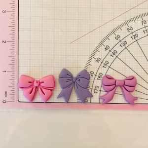 May include: Three decorative bows made of fondant, one pink, one purple, and one light pink. The bows are on a white surface with a ruler and protractor.