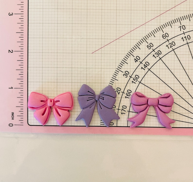 May include: Three decorative bows made of fondant, one pink, one purple, and one light pink. The bows are on a white surface with a ruler and protractor.