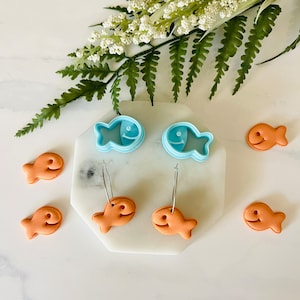 May include: Two pairs of orange fish-shaped earrings with silver hoops. The earrings are displayed on a white marble surface with two blue fish-shaped cookie cutters.