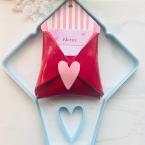 May include: A red envelope-shaped wall decoration with a pink heart and a light blue outline. The envelope contains a card with the word "Notes" and pink stripes. A second heart shape is cut out of the light blue outline.