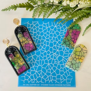 May include: A blue stencil with a stained glass window pattern. The stencil is used to create stained glass window designs in polymer clay. Two black and green stained glass window earrings are shown next to the stencil.