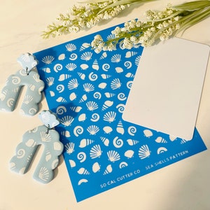 Puede incluir: Una plantilla azul con un patrón de conchas blancas. La plantilla está etiquetada como "So Cal Cutter Co. - Sea Shells Pattern". Se muestran dos pendientes de arcilla polimérica azul con un patrón de conchas junto a la plantilla.