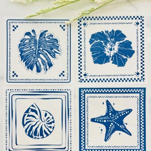 Puede incluir: Cuatro posavasos cuadrados con diseños azules y blancos. Cada posavasos presenta una imagen diferente: una hoja tropical, una flor, una concha de nautilus y una estrella de mar. Los posavasos tienen un borde decorativo.
