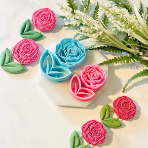 Op de afbeelding: Roze en blauwe oorbellen van polymeerklei in rozenvorm met bladeren. De oorbellen zijn verbonden met goudkleurige verbindingsringen. De afbeelding toont ook rozenvormige kleisnijders en kunstgroen.