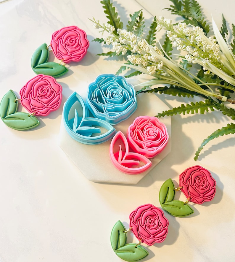 Op de afbeelding: Roze en blauwe oorbellen van polymeerklei in rozenvorm met bladeren. De oorbellen zijn verbonden met goudkleurige verbindingsringen. De afbeelding toont ook rozenvormige kleisnijders en kunstgroen.