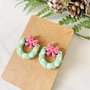 May include: A pair of green and white polka dot wreath earrings with pink bows. The earrings are on a brown card.
