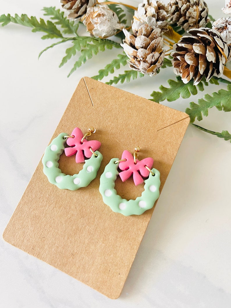 May include: A pair of green and white polka dot wreath earrings with pink bows. The earrings are on a brown card.