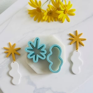 Pode incluir: Um conjunto de três brincos de argila polimérica com formato de nuvem branca, estrela amarela e flor azul. Os brincos são exibidos em uma superfície branca com um cortador de biscoitos em forma de flor azul e um cortador de biscoitos em forma de nuvem branca.