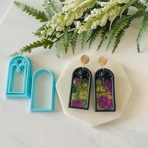 May include: A pair of black stained glass window earrings with gold hooks. The earrings are made of polymer clay and feature a colorful stained glass design. The earrings are shown next to two blue plastic cookie cutters shaped like stained glass windows.