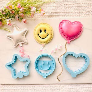 May include: Three balloon-shaped cookie cutters in light blue plastic, paired with three corresponding balloon-shaped cookies: a white star, a yellow smiley face, and a pink heart. Each cookie features a small ribbon or string detail.
