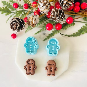 May include: Two blue gingerbread man cookie cutters and two brown gingerbread men cookies. The cookies are on a white marble surface. The cookie cutters are on top of the cookies.