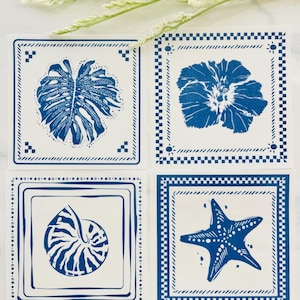 Pode incluir: Quatro bases de copos quadradas com desenhos azuis e brancos. Cada base de copo apresenta uma imagem diferente: uma folha tropical, uma flor, uma concha de nautilus e uma estrela do mar. As bases de copos t&ecirc;m uma borda decorativa.