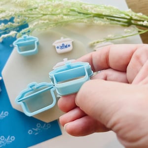 May include: Light blue plastic cookie cutters in the shape of a small casserole dish. Several cutters are visible, with one held in a hand. Some have a white and blue floral design. White flowers are in the background.