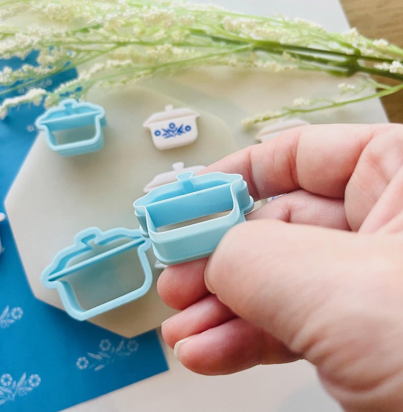May include: Light blue plastic cookie cutters in the shape of a small casserole dish. Several cutters are visible, with one held in a hand. Some have a white and blue floral design. White flowers are in the background.