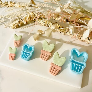 May include: A set of five cookie cutters in the shape of potted succulents. The cutters are light blue and the succulents are green. There are two small cutters, two large cutters, and one medium cutter.