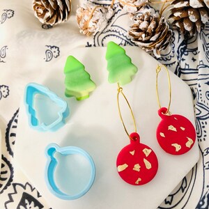 May include: A pair of gold hoop earrings with red and green Christmas ornament charms. The ornaments are decorated with gold flecks. The earrings are displayed on a white surface with a blue and white patterned fabric in the background.