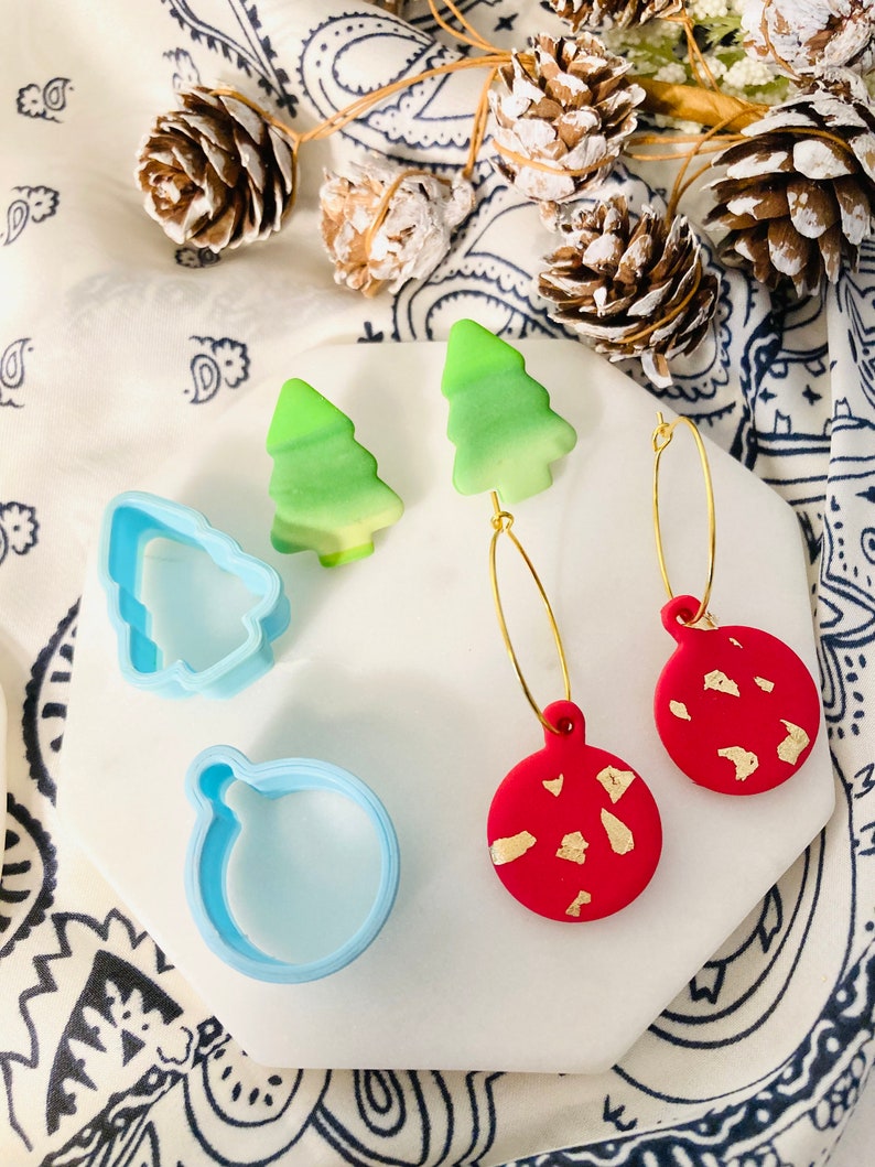 May include: A pair of gold hoop earrings with red and green Christmas ornament charms. The ornaments are decorated with gold flecks. The earrings are displayed on a white surface with a blue and white patterned fabric in the background.