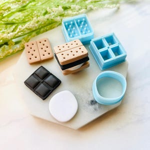 May include: An assortment of miniature polymer clay food items, including a s'more, chocolate square, and a white marshmallow. There are also light blue molds and a round cutter. The s'more is made of tan graham crackers, black chocolate, and white marshmallow.