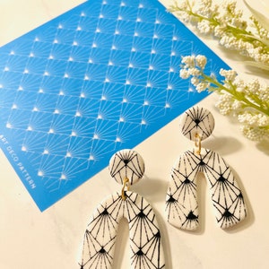 May include: A pair of white ceramic earrings with a black geometric pattern. The earrings are shaped like an arch and have a small round disc at the top. The earrings are on a white surface with a blue stencil that says "Art Deco Pattern".