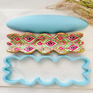 Puede incluir: Un cortador de galletas azul claro, una galleta decorada y un objeto ovalado azul claro. La galleta está decorada con un patrón verde, rosa y negro. El cortador de galletas tiene un borde ondulado.