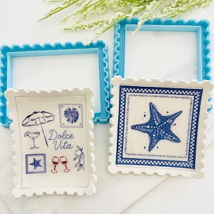 May include: Two white ceramic dishes with blue and red illustrations. One dish features a starfish design, the other has a cocktail umbrella, glasses, and the words "Dolce Vita". Two blue square frames are in the background.