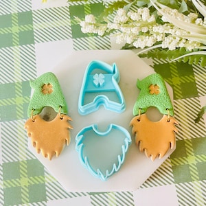 May include: A set of three blue plastic cookie cutters shaped like a gnome hat, beard, and a shamrock. The cutters are on a white surface with a green and white checkered background.