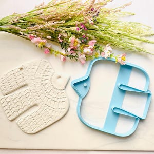 May include: A white ceramic bird-shaped ornament featuring an intricate embossed pattern of small arches and lines, paired with a light blue plastic cookie cutter in the same bird silhouette, positioned next to a bouquet of dried pink and green flowers.