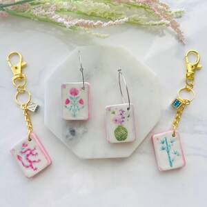 May include: Keychains and earrings featuring rectangular charms with floral and oriental designs. The charms have a white background with pink borders. The keychains have gold-colored clasps and dice charms. The earrings have silver hoops.
